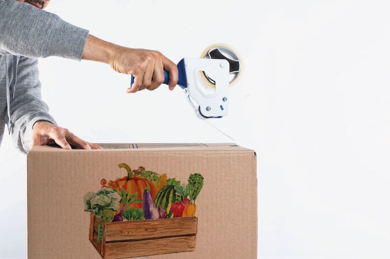 May include: A person is sealing a cardboard box with tape. The box has a colorful illustration of a wooden crate filled with fresh produce, including a pumpkin, eggplant, and various vegetables.