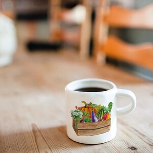 May include: White ceramic mug with a colorful illustration of a wooden crate filled with various vegetables and fruits. The mug is filled with coffee.