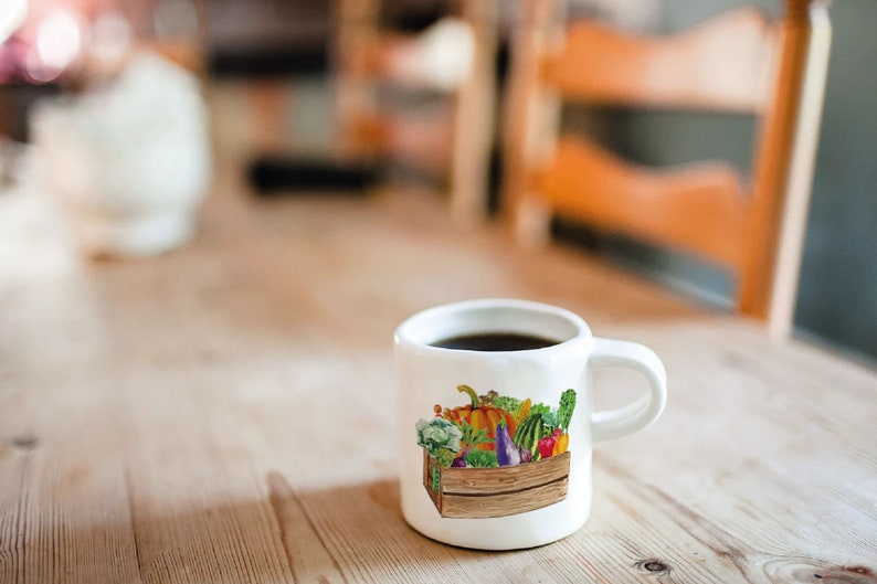May include: White ceramic mug with a colorful illustration of a wooden crate filled with various vegetables and fruits. The mug is filled with coffee.