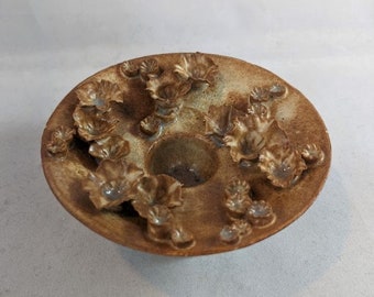 May include: A brown ceramic dish with a raised center and a ring of small, flower-shaped details around the edge.