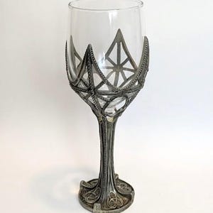 May include: A clear glass goblet featuring an intricate, dark metallic base and stem with a decorative lattice overlay that extends upward to cradle the bowl. The metalwork displays detailed geometric patterns and a textured, aged finish throughout.