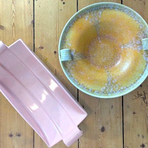 Puede incluir: Una bandeja rectangular de cerámica rosa con dos asas laterales y un cuenco redondo de cerámica amarilla con un esmalte blanco moteado y dos pequeñas asas laterales. Ambas piezas tienen un acabado esmaltado liso y formas funcionales.