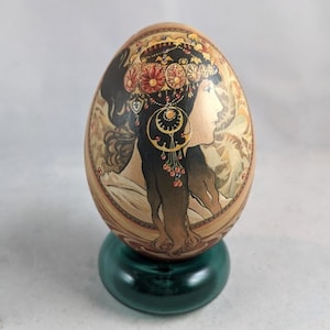 May include: A hand-painted egg featuring a detailed image of a woman with a floral crown and long hair. The egg is displayed on a green stand.