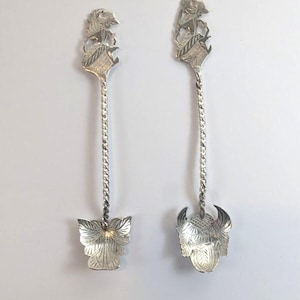 May include: Two ornate silver spoons. Each spoon has a detailed design at the handle's top and a floral or decorative element at the bowl. The handles have a twisted rope design.
