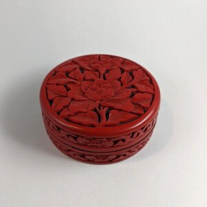 Puede incluir: Una caja de madera redonda de color rojo tallada con un diseño floral. La caja está cerrada y tiene una tapa.