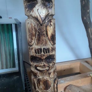May include: A carved wooden totem pole with a weathered appearance. The totem features a stylized face with large eyes and a prominent beak, along with other carved facial features. The wood has a natural, light brown color with darker, burnt accents.