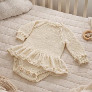 Könnte beinhalten: Cremefarbener gestrickter Baby-Strampler mit langen Ärmeln und Rüschenrock. Der Strampler hat dekorative Knöpfe und wird auf einer gesteppten Oberfläche zusammen mit einem Holzbeißring und einem wolkenförmigen Kissen präsentiert. Gemütliches, neutrales Kinderzimmer-Thema.