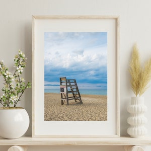 May include: A framed photograph of a wooden lifeguard stand on a sandy beach with a blue sky and white clouds. The stand has a sign that reads "Lifeguard on Duty".
