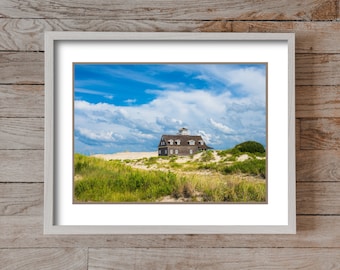 May include: A gray wooden house with white trim sits on a sand dune with green grass and a blue sky with white clouds.