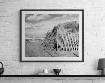 May include: Black and white photograph of a wooden fence on a sandy beach. The fence is partially collapsed and the sand is blowing in the wind. The ocean is in the background.