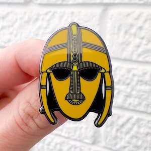 May include: A yellow enamel pin featuring a stylized face wearing a helmet with intricate details. The helmet has a black outline and the face has black eyes and a black mouth.