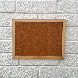 Puede incluir: Un corcho rectangular con una superficie marrón claro y un marco de madera natural. El corcho está montado en una pared de ladrillo blanco. La imagen es un plano medio, que muestra el corcho centrado.