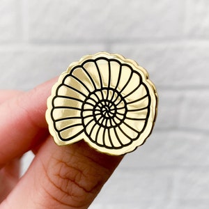 May include: Gold-colored pin with a black outline of a seashell. The seashell has a spiral pattern.