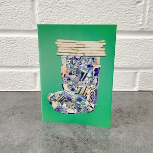 以下が含まれることがあります： 青と白の陶器の破片で作られたブーツのデザインが施された緑色のグリーティングカード。ブーツの上部には、小さな長方形のピースが飾られています。カードは灰色の表面に置かれ、背景には白いレンガの壁があります。