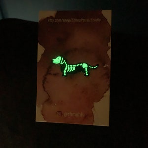 May include: A glow-in-the-dark dachshund skeleton enamel pin. The pin is on a white card with watercolor-style pink and brown splotches. The card also includes the text "etsy.com/shop/EmmaYouellStudio" and "@ehmahh".