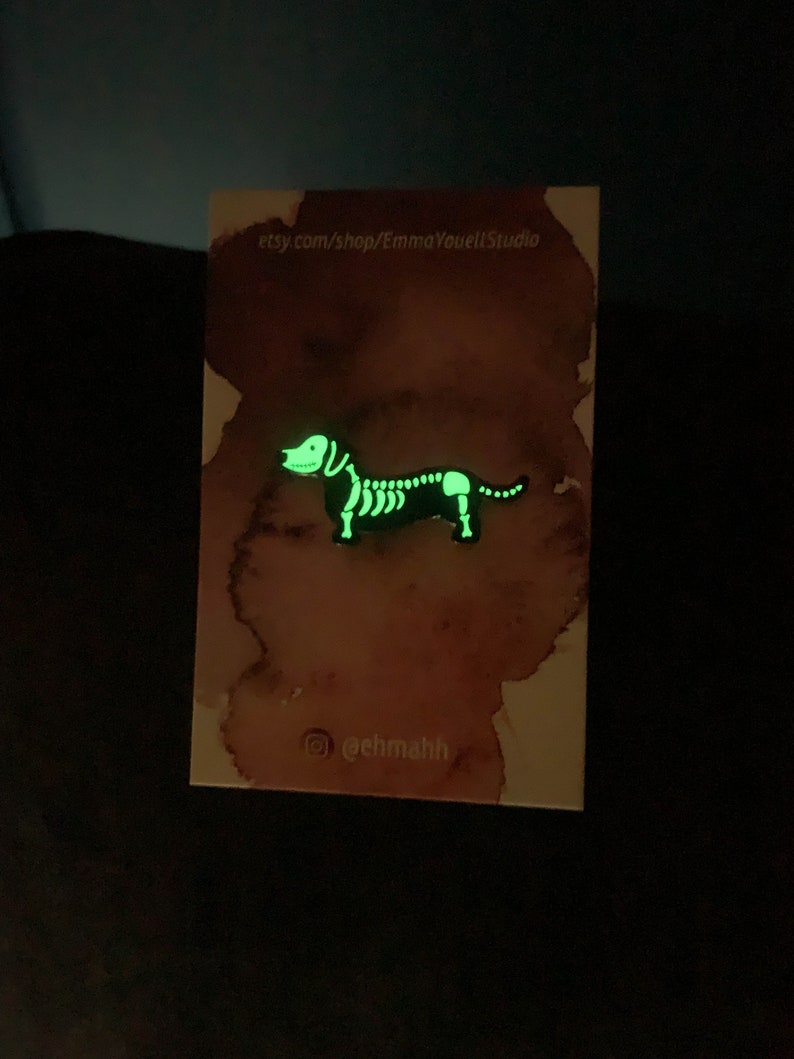 May include: A glow-in-the-dark dachshund skeleton enamel pin. The pin is on a white card with watercolor-style pink and brown splotches. The card also includes the text "etsy.com/shop/EmmaYouellStudio" and "@ehmahh".