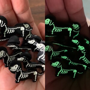 May include: A set of glow-in-the-dark dachshund skeleton pins. The pins are black with white skeleton details and glow green in the dark.
