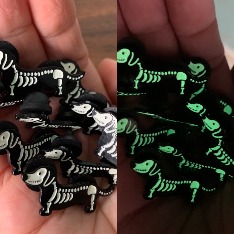May include: A set of glow-in-the-dark dachshund skeleton pins. The pins are black with white skeleton details and glow green in the dark.
