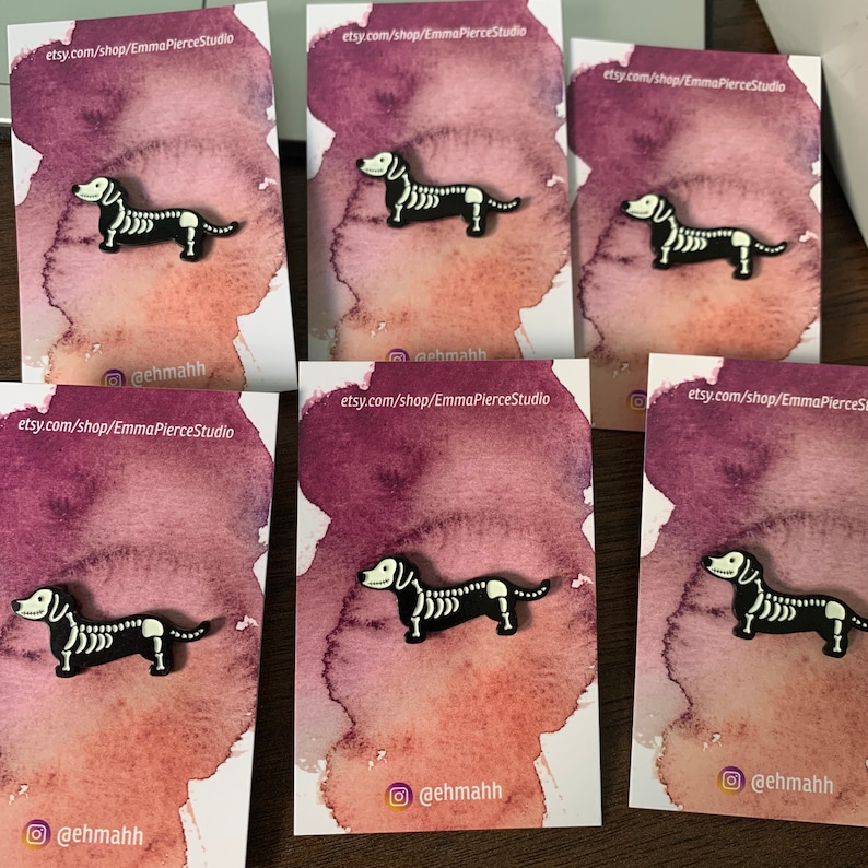 May include: Five cards with a black and white skeleton dachshund pin on each. The cards have a watercolor background with a pink and purple gradient. The text on the cards reads "etsy.com/shop/EmmaPierceStudio" and "@ehmahh".