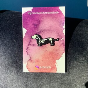 May include: A black and white enamel pin of a dachshund skeleton on a pink and purple watercolor background. The pin is on a white card with the text "etsy.com/shop/EmmaYouellStudio" and "@ehmahh".