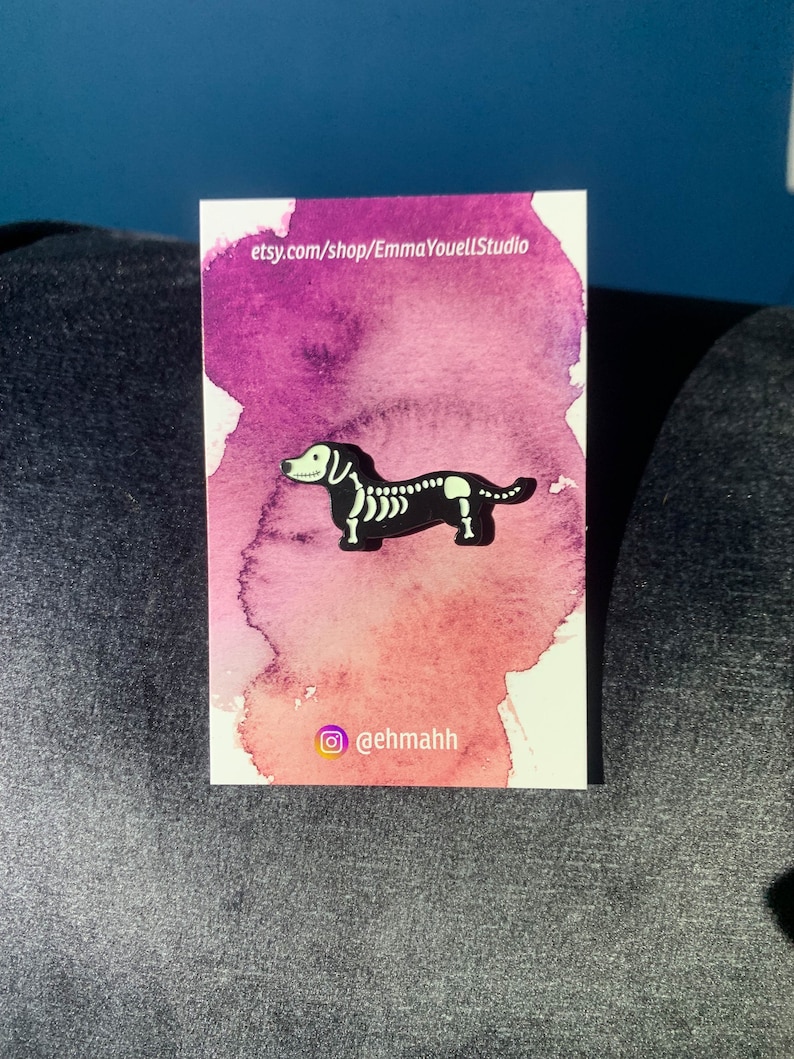 May include: A black and white enamel pin of a dachshund skeleton on a pink and purple watercolor background. The pin is on a white card with the text "etsy.com/shop/EmmaYouellStudio" and "@ehmahh".