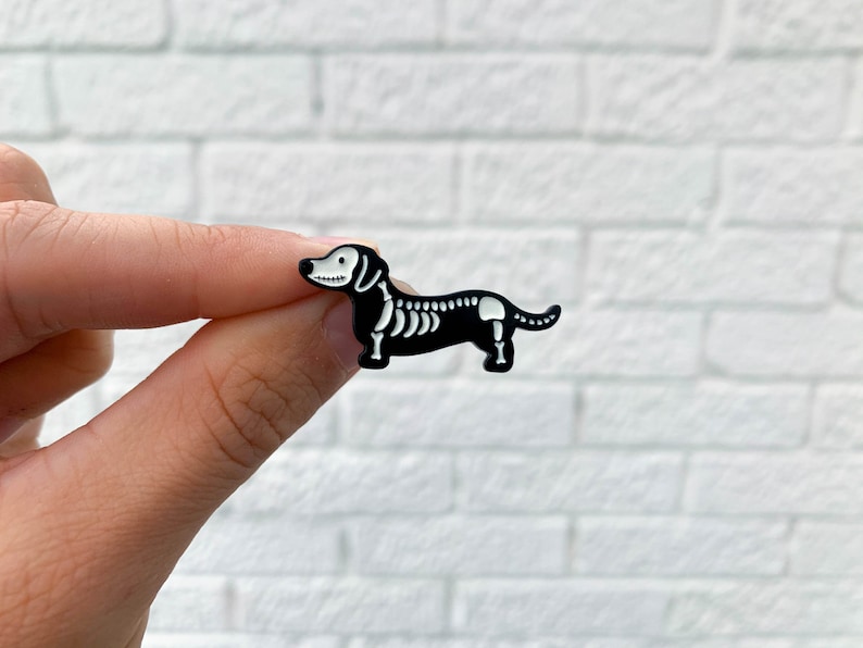 A hand holds an enamel lapel pin. The pin is shaped like a dog. A miniature dachshund enamel pin in black that has glow-in-the-dark bones.