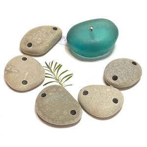 May include: Six smooth, gray stones with two holes drilled through each. The stones are arranged in a circle around a teal blue glass disc.
