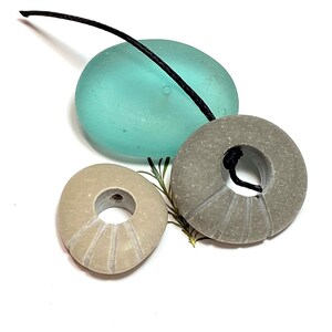 May include: Three round stone beads with holes drilled through the center. The beads are light brown and gray with a textured surface. A black cord is threaded through the beads.