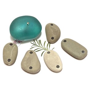 May include: Five smooth, gray stones with two holes drilled through each. The stones are arranged on a white surface with a sprig of green foliage and a turquoise glass sphere.