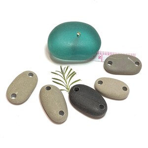 May include: Five smooth, oval-shaped stones with two holes drilled through each. The stones are in various shades of gray and brown. A teal-colored glass object is in the background.