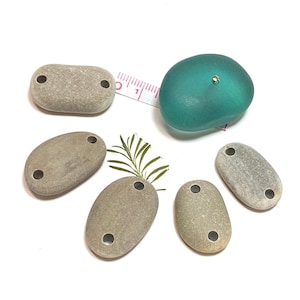 May include: Six smooth, oval-shaped stones with two holes drilled through each. The stones are a light brown color and are arranged in a circle around a teal-colored glass bead.
