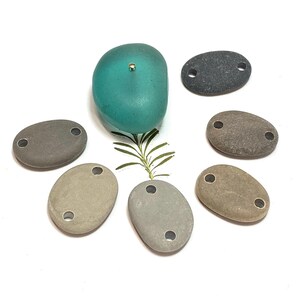 May include: Seven smooth, oval-shaped stones with two holes drilled through each. The stones are in various shades of gray and brown. A teal-colored glass stone with a gold pin in the center is in the middle of the arrangement.