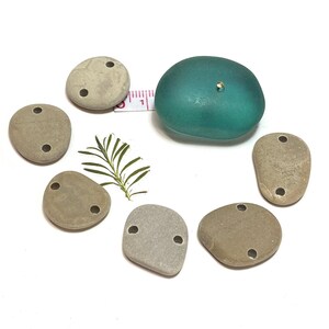 May include: Seven smooth, gray stones with two holes each. The stones are arranged in a circle around a teal-colored glass object with a gold accent.