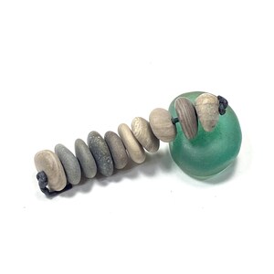 May include: A string of ten smooth, rounded, multi-colored gray and beige stones threaded onto a dark cord, positioned next to a single, translucent, rounded piece of sea-foam green glass that has a smooth, polished surface texture.