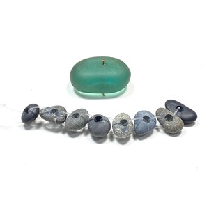 May include: A single smooth, translucent teal sea glass piece with a small metal pin inserted at the top, positioned above a string of eight varied gray and dark blue natural stones, each featuring a central hole for threading on a clear cord.