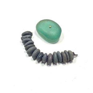 May include: A collection of twelve small, smooth, dark gray stones strung together in a curved line, accompanied by a single, larger, translucent sea-foam green glass pebble featuring a small, centered gold-colored metal stud.