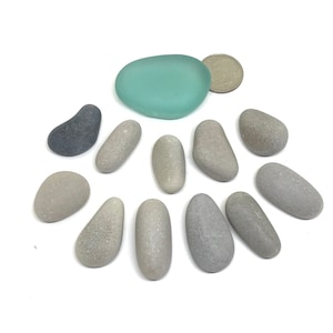 May include: A collection of smooth, gray and light blue sea glass stones. The stones are arranged in a circle on a white background.