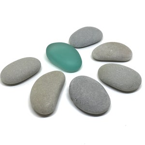 May include: A collection of seven smooth stones, six gray and one light blue, arranged in a circle on a white background.