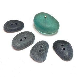 May include: A collection of five buttons in various shapes and colors. Four are oval-shaped, dark gray with two holes each. One is a larger, oval-shaped, translucent teal button with a small gold-colored embellishment.