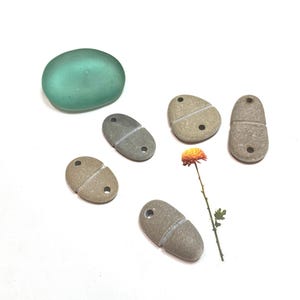 May include: An assortment of small, gray stone pendants with a split design and drilled holes, alongside a smooth, sea-green glass stone. A single orange flower adds a touch of color to the composition. The pendants are likely for jewelry making.