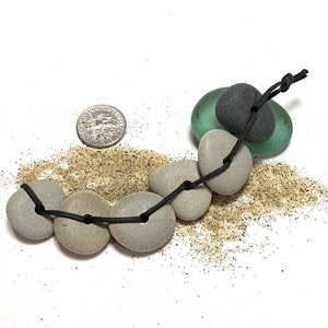 May include: A necklace made of smooth, gray stones and green and black glass beads. The stones are strung on a black cord and tied together with a knot. The necklace is surrounded by sand.