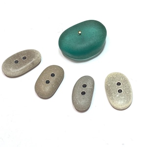 May include: A collection of handmade buttons in various shades of gray and a teal green. The buttons are oval-shaped with two holes each, resembling small pebbles. The teal button has a gold accent.