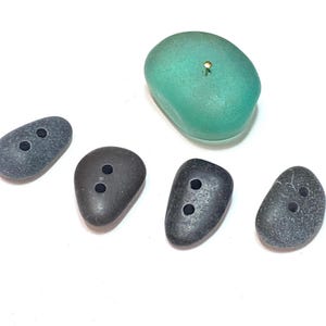 May include: A collection of stone-shaped buttons in various shades of gray and a single teal-colored button with a gold-colored pin. The buttons have two holes each for sewing onto fabric. The buttons are arranged on a white surface.