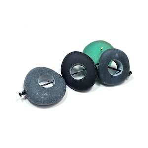 May include: Three smooth, rounded stones with central holes, strung together on a thin black cord. Two stones are dark gray, and one is solid black. A small, rounded green object is positioned behind the black stone, partially visible.