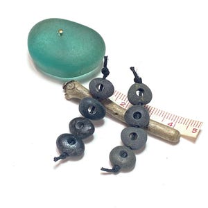 May include: An arrangement of natural elements. A smooth, oval, seafoam green stone with a small gold-colored pin is at the top. Below are several dark gray, circular beads strung on black cord. A small wooden stick and a ruler are also present.