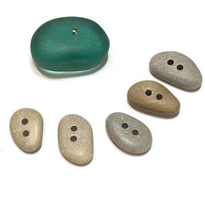 May include: A collection of unique buttons in various shapes and colors. One large, teal-colored, rounded button with a gold-colored pin is at the top. Below are six smaller, pebble-shaped buttons in shades of gray and tan, each with two holes.