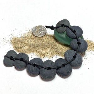 May include: A black cord bracelet with smooth, dark gray stones. The stones are strung together in a circular pattern and are tied with a black cord. The bracelet is lying on a white surface with a small pile of sand.