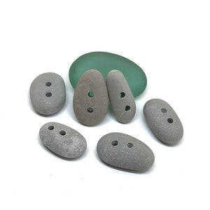 May include: Six gray oval-shaped stones with two holes each. The stones are arranged in a semi-circle on a white surface. A piece of green glass is behind the stones.