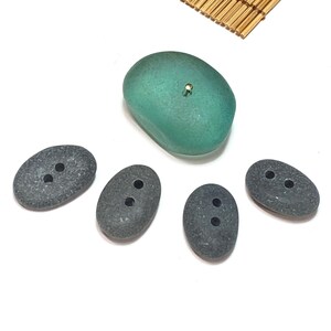 May include: An assortment of stone buttons and a larger teal-colored stone with a gold-colored pin. The buttons are oval-shaped, gray, and have two holes. The teal stone is smooth and rounded.