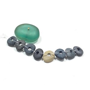 May include: A strand of eight small, dark gray and tan stones with center holes threaded on a clear filament, accompanied by a single, smooth, translucent sea-glass green oval stone featuring a small gold-colored metal pin at the top.